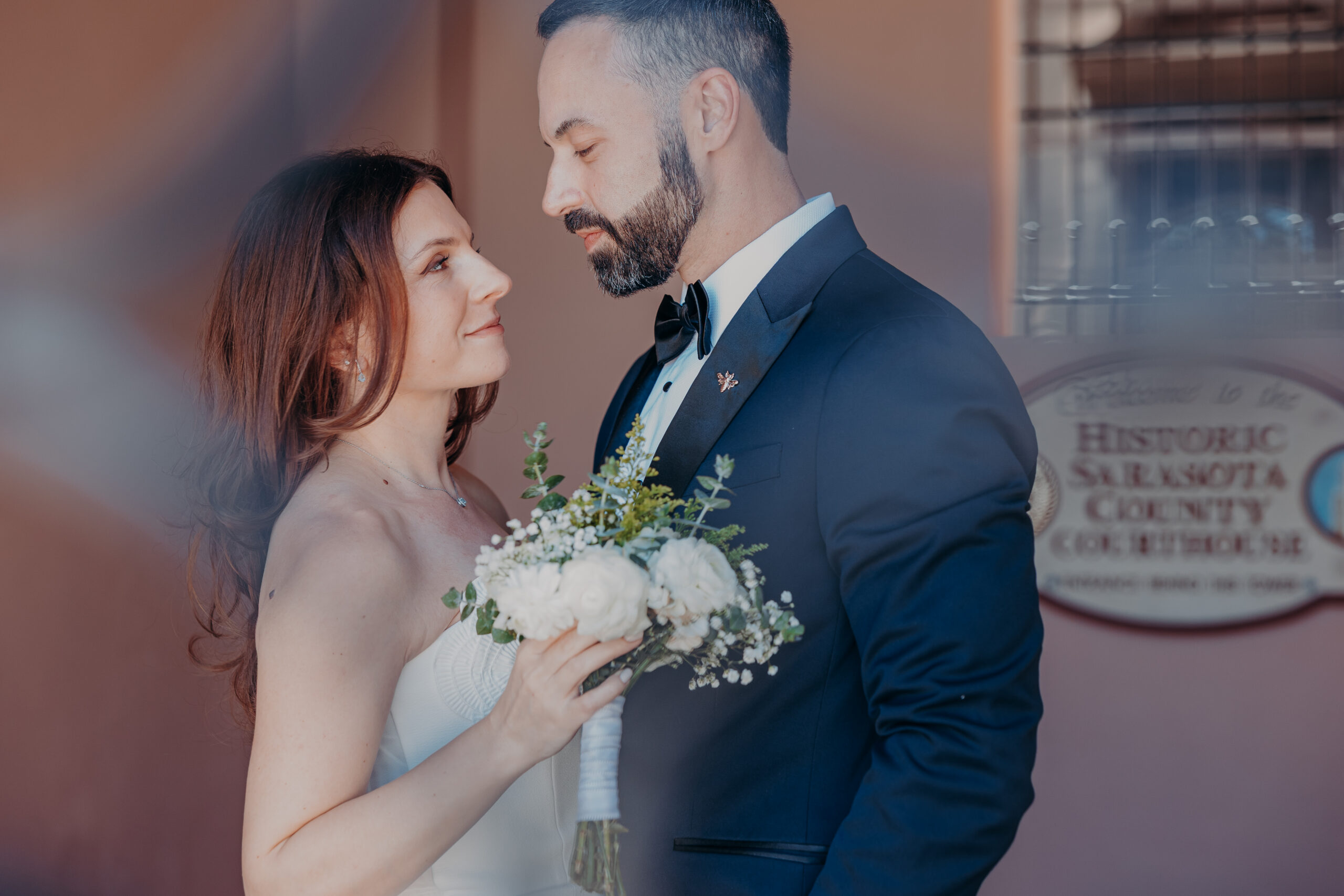 Diana and Robert gazing at each other in front of the Historic Sarasota County Courthouse sign during their wedding portraits