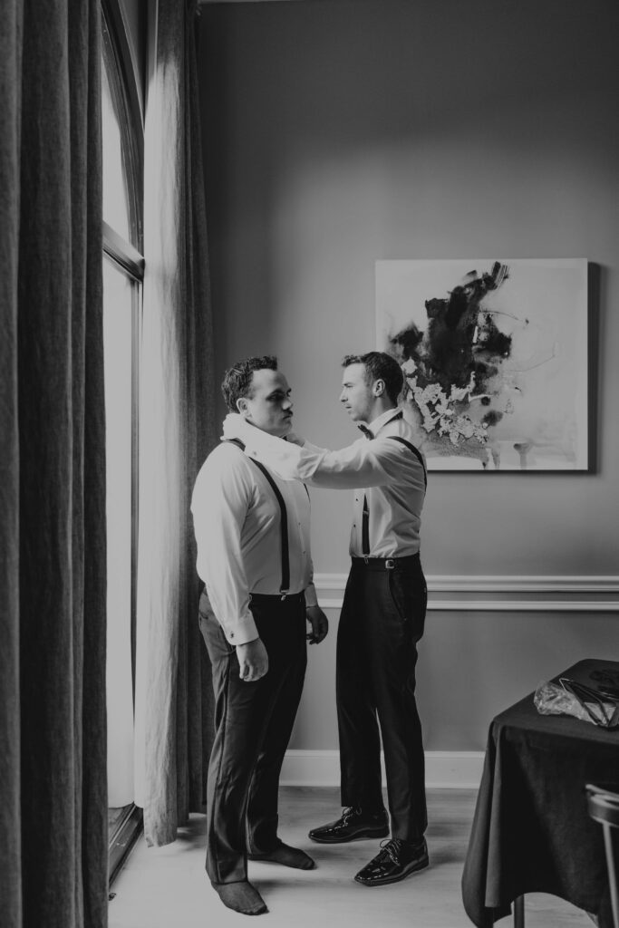 Black and white groom and groomsman in suspenders getting ready by window at Lakewood Ranch wedding
