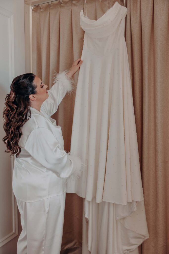 Bride in white feather-trimmed robe reaching up to touch her pearl-dotted wedding gown