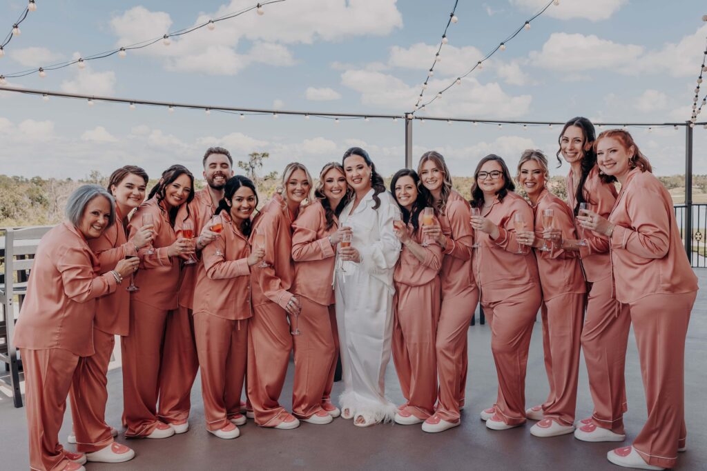 Bride surrounded by bridesmaids in dusty rose satin pajamas holding champagne on rooftop terrace