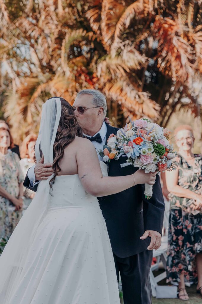 Father of the bride kissing his daughter on cheek at ceremony altar with colorful bouquet and palms