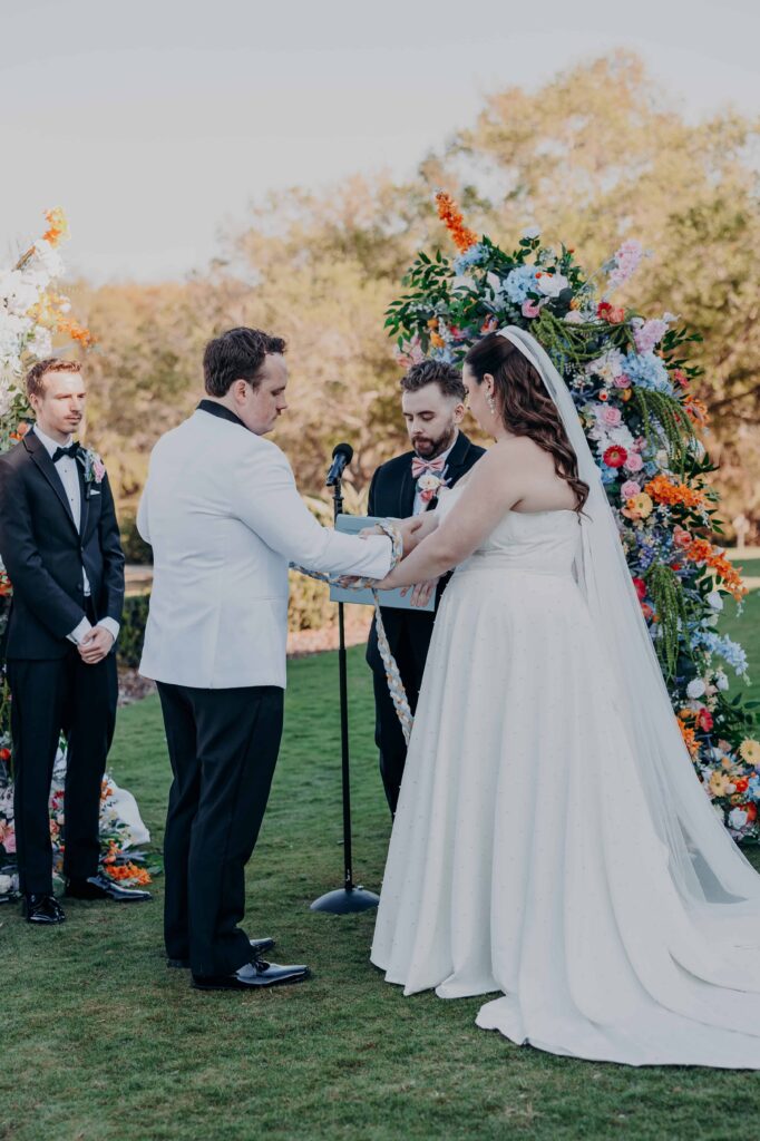 Handfasting ceremony with hands bound by braided blue gold and white cords in front of colorful floral arch